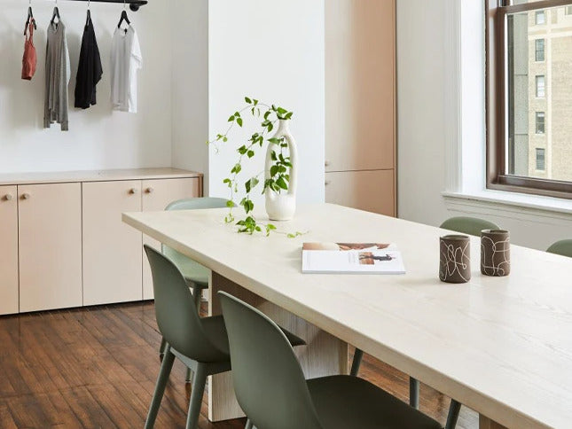 Photo of our bright white conference room in our office with a large rectangular table as the focal point, two sage green chairs tucked in, a window viewing NYC buildings at the back, a plant and book on the table, and four styles hanging at the back rack